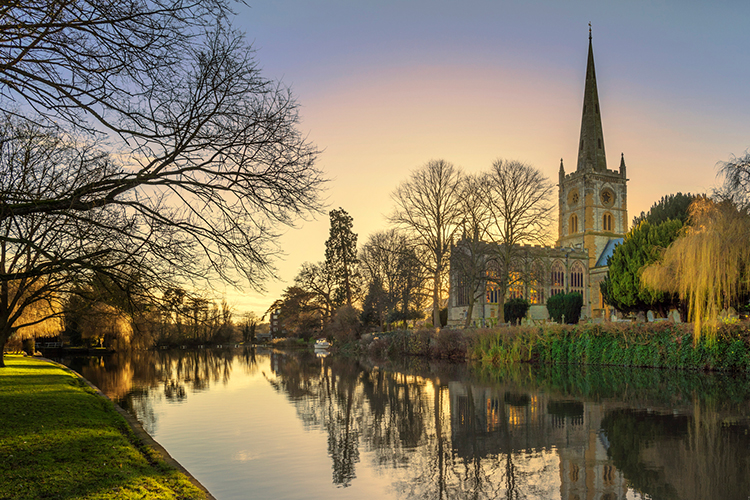 A village in Warwickshire that shows a river and a church on the right hand side of it. style=